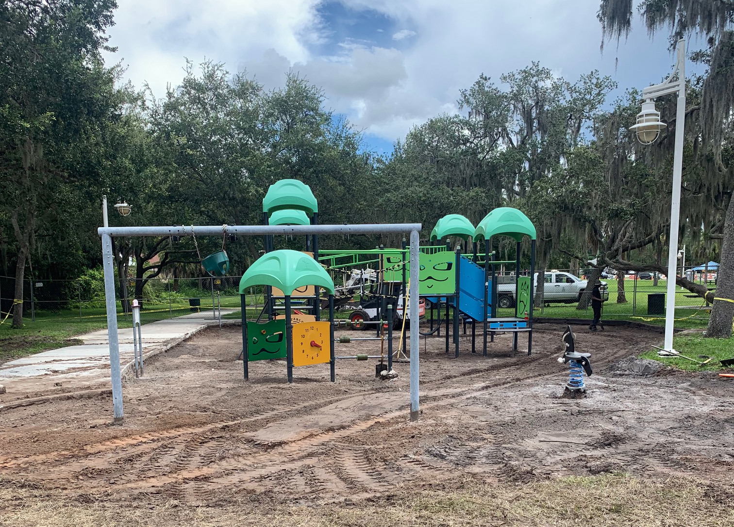 Playground with surface removed on the ground with dirt. Playground is yellow blue green and black with a slide and swings on a cloudy day.